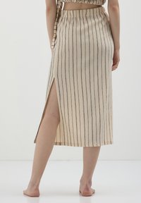 Femme portant une jupe midi à rayures verticales beige et noire avec une fente sur le côté, debout pieds nus sur un sol blanc contre un fond uni.