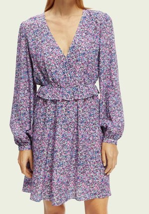Femme portant une robe longue à manches, fleurie rose et violette avec un col en V et une taille cintrée, debout devant un fond uni.