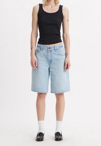 Henkilö, jolla on päällään musta hihaton toppi, vaaleansiniset denim-Shortsit polviin asti, valkoiset sukat ja mustat loafersit, seisoo valkoista taustaa vasten.