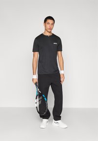 Melna sporta t-krekls ar "BOSS" logotipu, saskaņots ar melnām sporta biksēm. Modelis tur tenisa raketi un valkā baltas sporta kurpes.