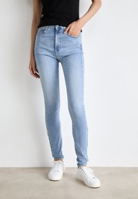 Jeans ajustados de talle alto azul claro, hechos de denim con una textura suave. Presentan cinco bolsillos y una cremallera, combinados con zapatillas blancas.