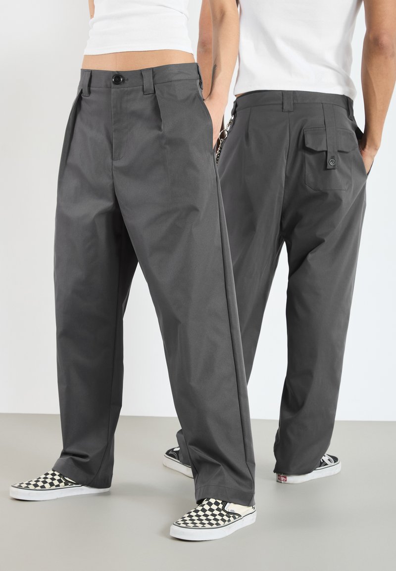 Graue plissierte Hose mit einem Frontknopfverschluss, seitlichen Taschen und einer Cargo-Tasche an der Rückseite, kombiniert mit schwarz-weißen, karierten Slippern.