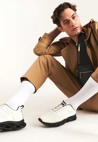 Sneakers bianchi da uomo con suola nera, materiale liscio, lacci anteriori. L'outfit presenta una giacca beige e pantaloni slim-fit, abbinati a calze bianche.