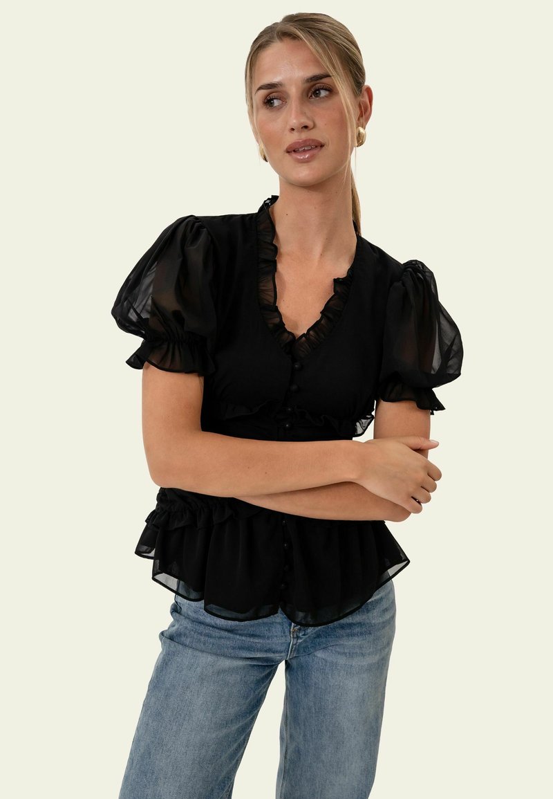Femme portant un chemisier noir à volants avec des manches bouffantes et un jean bleu, bras croisés, regardant vers sa gauche sur un fond clair.