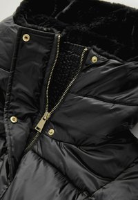Jachetă puffer neagră cu guler din blană sintetică, fermoar aurit și închideri cu capse. Captușeală interioară texturată și model matlasat pe exterior.