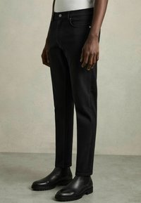 Jeans negre din denim cu o croială slim, dotate cu cinci buzunare, închidere frontală cu nasture și detalii de cusătură subtile, asortate cu bote negre din piele până la glezne.