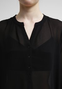 Žena má na sobě černou průhlednou blůzu s výstřihem do V a třemi černými knoflíky vpředu, přes černý tílko.