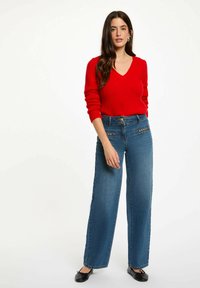 Maglione rosso a scollo a V abbinato a jeans blue a vita alta e gamba larga con tasche decorative sul davanti. Indossato con ballerine nere. Sfondo semplice.