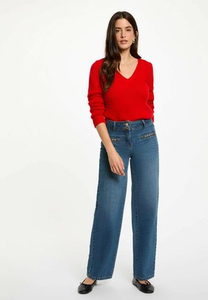 Maglione rosso a scollo a V abbinato a jeans blue a vita alta e gamba larga con tasche decorative sul davanti. Indossato con ballerine nere. Sfondo semplice.