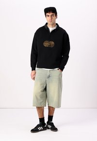 Sudadera negra con bordado dorado, cuello con media cremallera, combinada con pantalones cortos claros desteñidos y zapatillas negras. Diseño minimalista en exhibición.