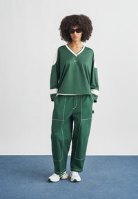 Osoba s kučeravými vlasmi, ktorá má na sebe tmavé slnečné okuliare, zelenú dlhú košeľu s bielymi detailmi, voľné zelené nohavice a biele tenisky, stojí na modrej podlahe.