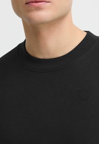 Zwarte T-shirt met een ronde hals, gemaakt van zachte katoen. Heeft een subtiele reliëflogo aan de onderlinkerkant.