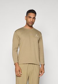 Long-sleeve, beige t-shirt made of soft fabric, featuring a rounded neckline and small white logo on the left chest area.