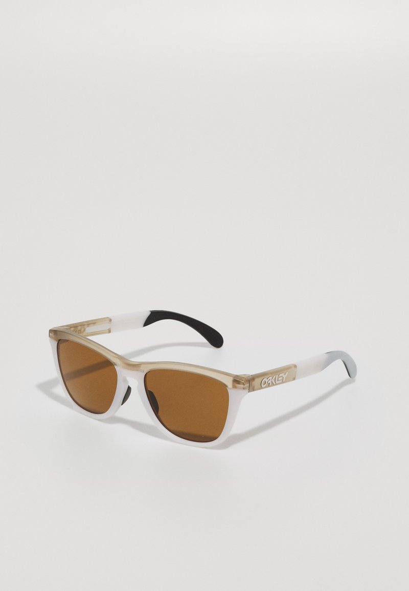 Ochelari de soare cu nuanță maro, cu un cadru transparent de culoare bej, cu vârfuri negre la brațe și un logo argintiu pe lateral; design unghiular și dimensiune medie.