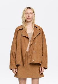 Bruine suède oversized jas met een brede kraag en knoopsluiting, gecombineerd met een bijpassende suède minirok. Soepele textuur, minimalistisch design.