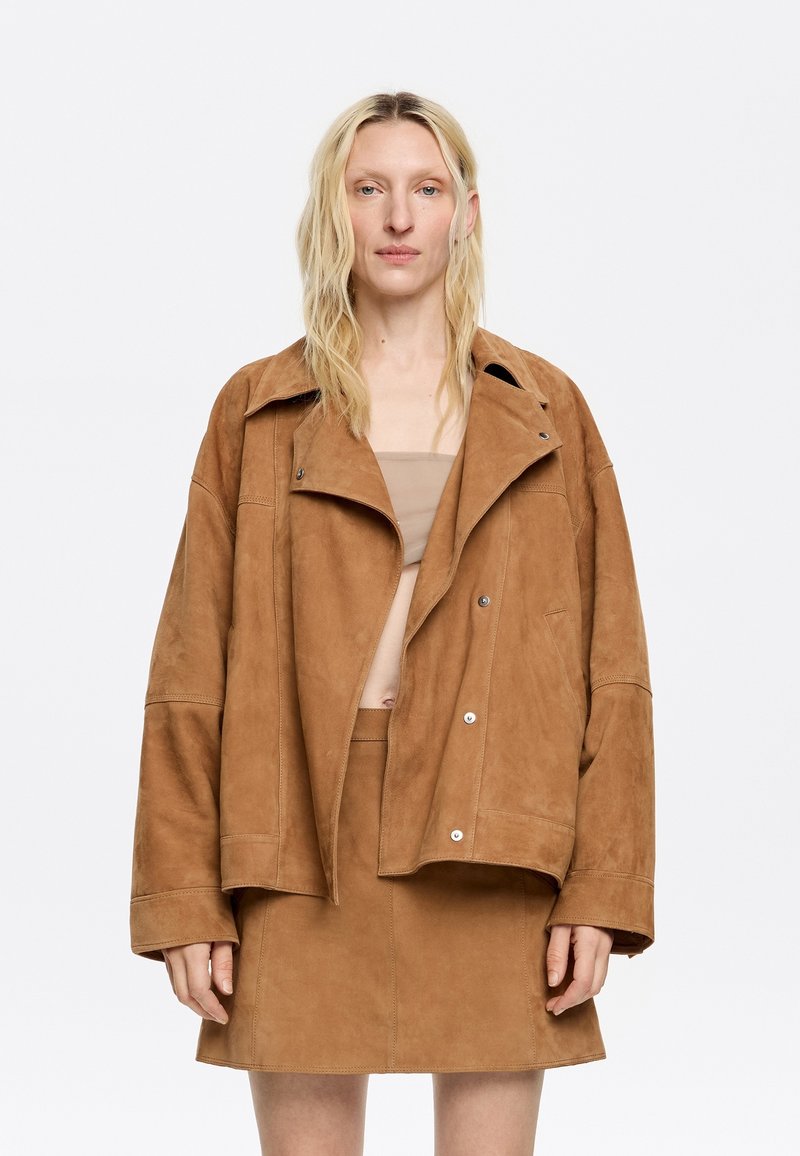 Bruine suède oversized jas met een brede kraag en knoopsluiting, gecombineerd met een bijpassende suède minirok. Soepele textuur, minimalistisch design.