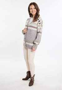 Cardigan grigio a maglia con motivi geometrici in crema, marrone e grigio scuro; maniche lunghe, chiusura a bottoni, indossato sopra una maglietta chiara e pantaloni beige.