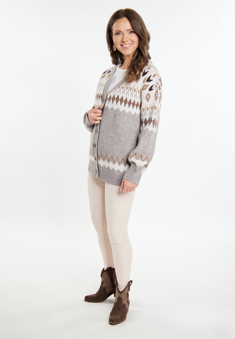 Grå stickad cardigan med geometriska mönster i kräm, brunt och mörkgrått; långa ärmar, knäppning framtill, bärs över en ljus topp och beige byxor.