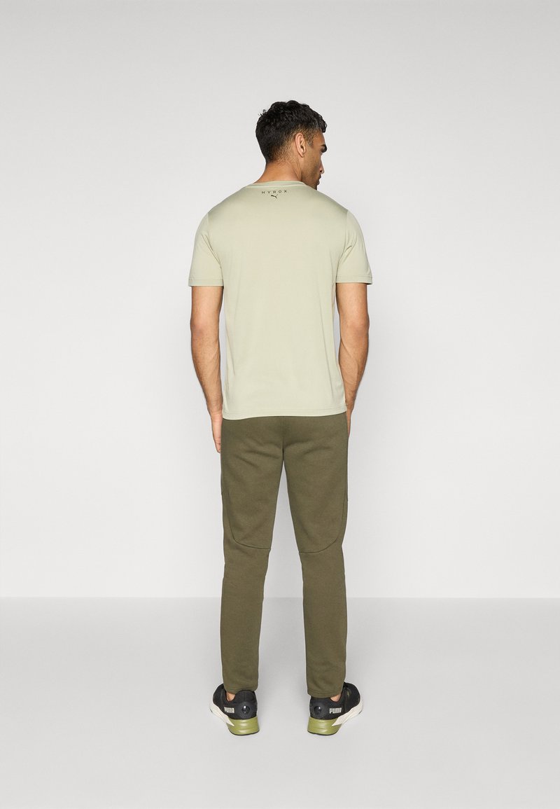T-shirt vert clair à manches courtes avec un col rond et un logo dans le dos, associé à un pantalon vert olive et des baskets noires.