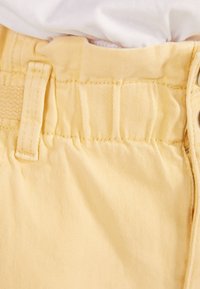 Gula bomullsshorts med texturerad tyg, med en rynkad midja och bälteshål, närbild av den övre sektionen.