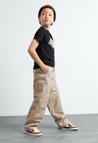 T-shirt graphique noir avec logo, pantalon cargo beige avec plusieurs poches, et baskets marron avec lacets blancs. L'enfant pose les mains dans les poches.