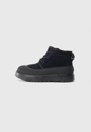 Chaussure noire montante jusqu'à la cheville avec semelle en caoutchouc épaisse, tige en daim, fermeture lacée à l'avant, et capuchon de orteil texturé, présentée en profil latéral sur fond blanc.