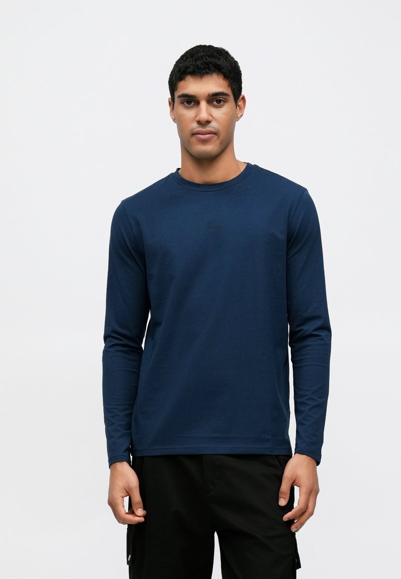 Μακρυμάνικη μπλούζα σε ναυτικό μπλε χρώμα από βαμβάκι με στρογγυλή λαιμόκοψη και διακριτικό λογότυπο της μάρκας στο στήθος, συνδυασμένη με μαύρο παντελόνι.