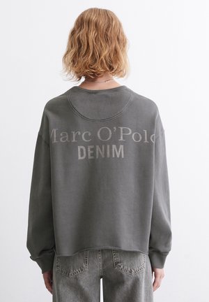 Person med kort bølget blondt hår iført en grå oversized sweatshirt med teksten "Marc O'Polo DENIM" og grå denimjeans, set bagfra.