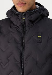 Giubbotto imbottito nero con un motivo a zigzag testurizzato, colletto alto, zip frontale e patch con logo. Il materiale sembra isolante e leggero.