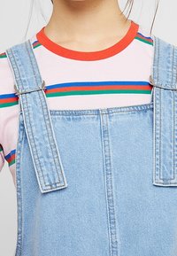 Ljust blå denimoverall med silverdetaljer, bärs över en rosa randig t-shirt med röda, blå och gröna horisontella linjer.