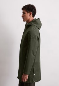 Mężczyzna zwrócony w lewo, ubrany w ciemnozieloną parkę z kapturem, posiadającą boczne kieszenie i zatrzaski na dolnym brzegu, na tle jednolitego, jasnego tła.