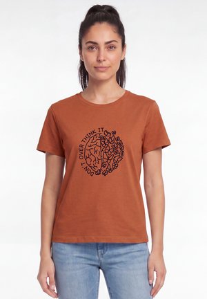 Vrouw draagt een roestkleurig T-shirt met een afbeelding van een brein en vlinders en tekst "Don't over think it", staand tegen een effen witte achtergrond.
