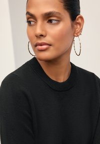 Maglione nero lavorato a maglia con scollo rotondo, abbinato a grandi orecchini a cerchio dorati in stile bambù. Texture liscia, primo piano di volto e parte superiore del corpo.