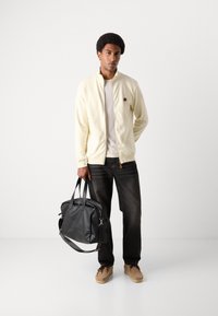 Lys gul zip-up jakke med tekstureret stof, grå t-shirt, sorte jeans, beige ruskindssko og en stor sort lædertaske.