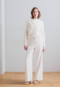 Hvit teksturert cardigan med gullknapper, kombinert med hvite vide bukser. Modellen står mot en lysegrå vegg på tregulv.