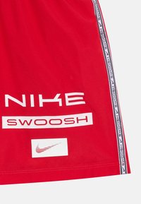 Röda atletiska shorts med vit text "NIKE" och "SWOOSH". Sidsnöret visar upprepande svart bokstäver och den ikoniska Nike-logotypen. Slät tygtextur.