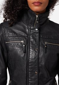Schwarze Lederjacke mit hohem Kragen, Reißverschlusstaschen und strukturierten Einsätzen auf den Schultern. Glatte Oberfläche und glänzende Hardware-Details.