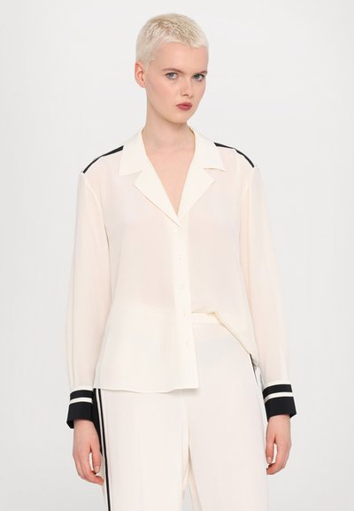 Blusa crema con vestibilità rilassata, chiusura con bottoni e polsini a strisce nere a contrasto. Tessuto leggero e testurizzato con colletto.