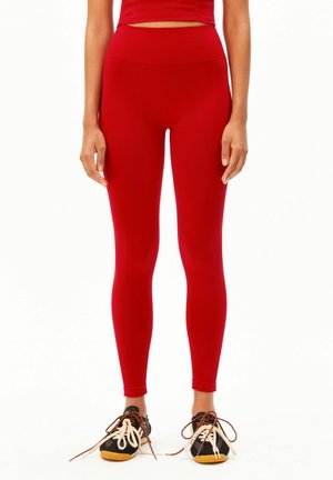 Vrouw draagt rode leggings met hoge taille en zwarte sneakers met grote, niet-overeenkomende veters, staand tegen een witte achtergrond.