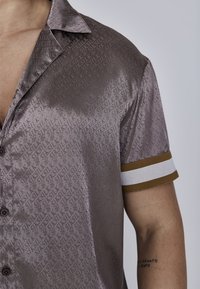 Camisa de botón de manga corta en una suave y brillante tela malva, que presenta un sutil patrón de logotipos en toda la prenda y puños a rayas en marrón y blanco.