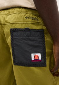 Μαύρη τσέπη με λογότυπο σιλουέτας ψαρέματος ραμμένη σε ελιά-πράσινο σορτς Columbia Pro, που φοριέται από άτομο με σκούρο δέρμα.