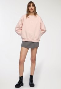 Różowy sweter z napisem "SELFLOVE" w kolorze białym. Połączony z szaro-plecioną mini spódnicą i czarnymi, sznurowanymi botkami. Jednolity tło.