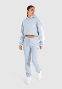 Sudadera corta de color azul claro con mangas y logo blancos, combinada con pantalones de chándal azules a juego. Tela suave con un ajuste relajado. Tenis blancos.