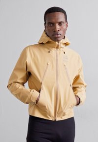 Veste imperméable jaune avec capuche, présentant des poches avant zippées et une fermeture éclair à l'avant. Matière lisse et légère.