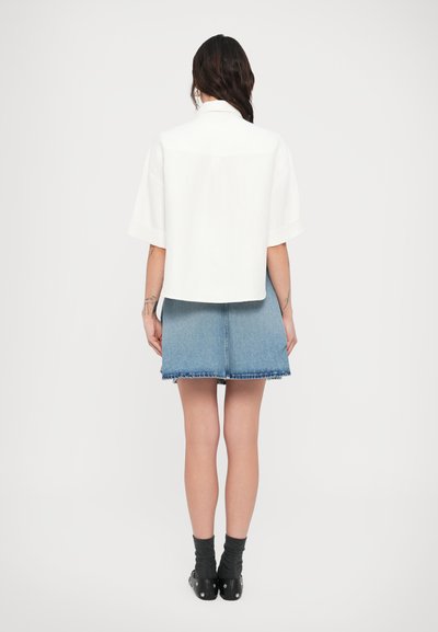 Λευκό πουκάμισο με κοντό μανίκι και γιακά, συνδυασμένο με φούστα από γαλάζιο τζιν με ακατέργαστο τελείωμα. Μαύρα μποτάκια αστραγάλου και γκρι κάλτσες.