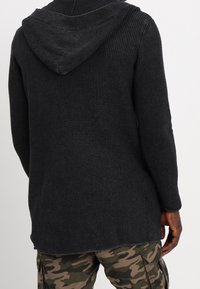 Hoodie noir texturé avec un motif côtelé, doté d'une capuche et d'une coupe décontractée. Porté avec un pantalon cargo camouflage vert.