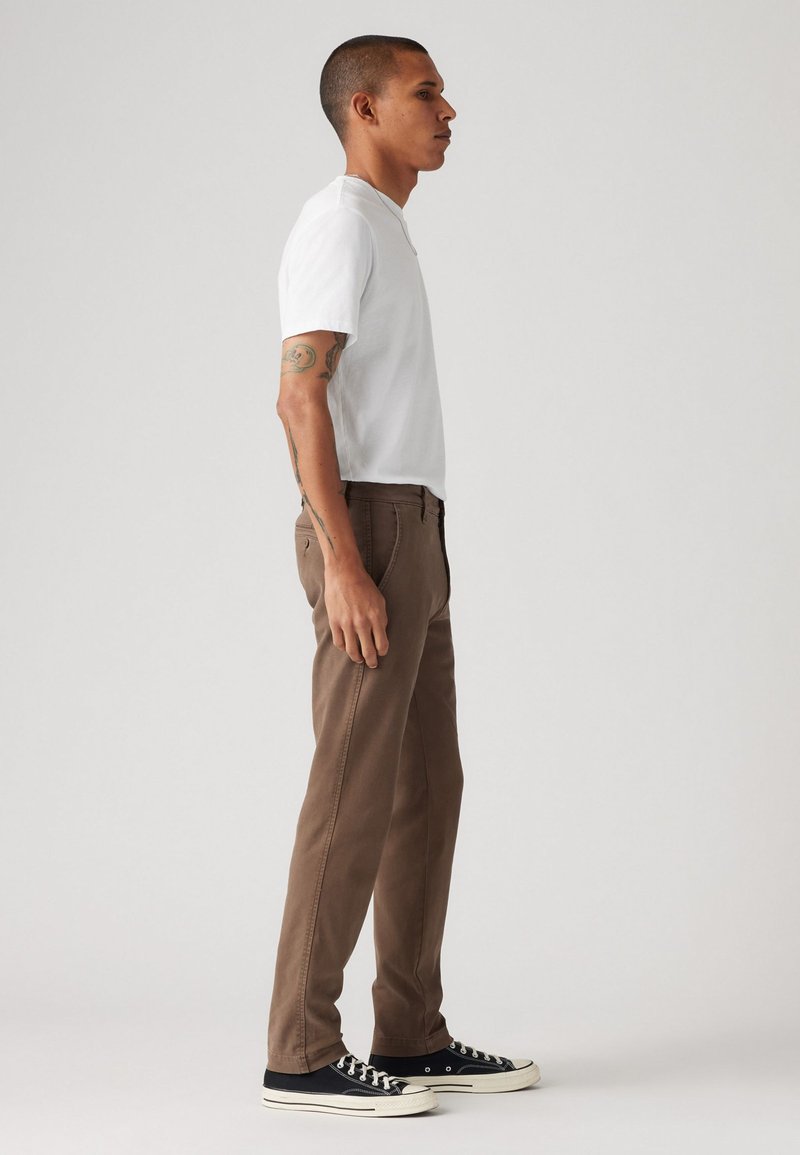 Pantalons en coton marron avec une coupe droite, associés à un t-shirt blanc à manches courtes et des baskets basses noires. Aucun motif ou accent notable.