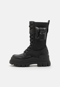 Bottine de combat noire à lacets fabriquée en matériau synthétique. Elle présente un dessus texturé, une semelle épaisse et une accentuation par boucle latérale pour une fermeture sécurisée.