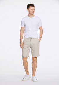 Shorts en lin gris clair avec une taille à cordon, associés à un t-shirt blanc ajusté et des baskets blanches, se tenant devant un fond uni.