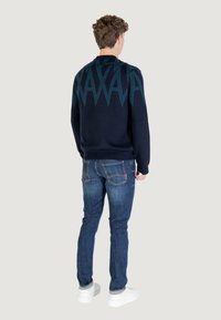 Homme de dos portant un pull bleu marine à motif géométrique, un jean bleu et des baskets blanches sur un fond uni.